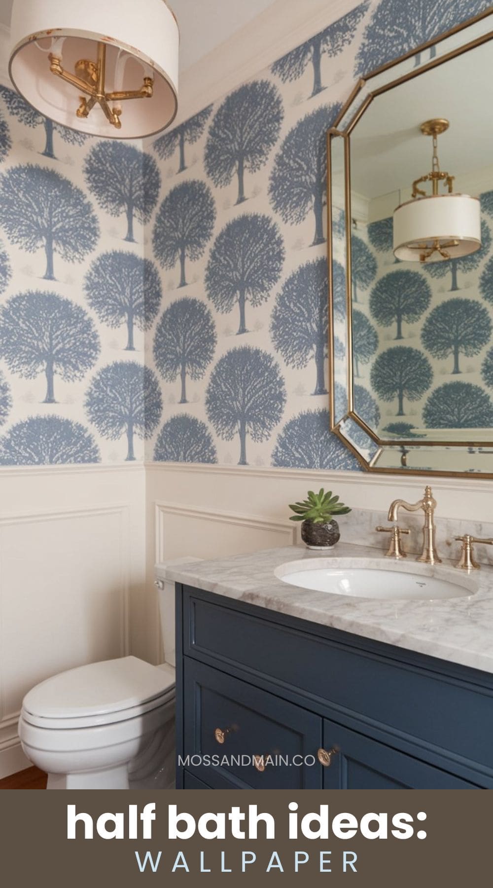 A stylish half bath features tree-patterned wallpaper, a large rectangular mirror with a gold frame, a marble countertop, and a small succulent. Theres a blue vanity with brass fixtures and a white toilet. The ceiling light has a beige shade.