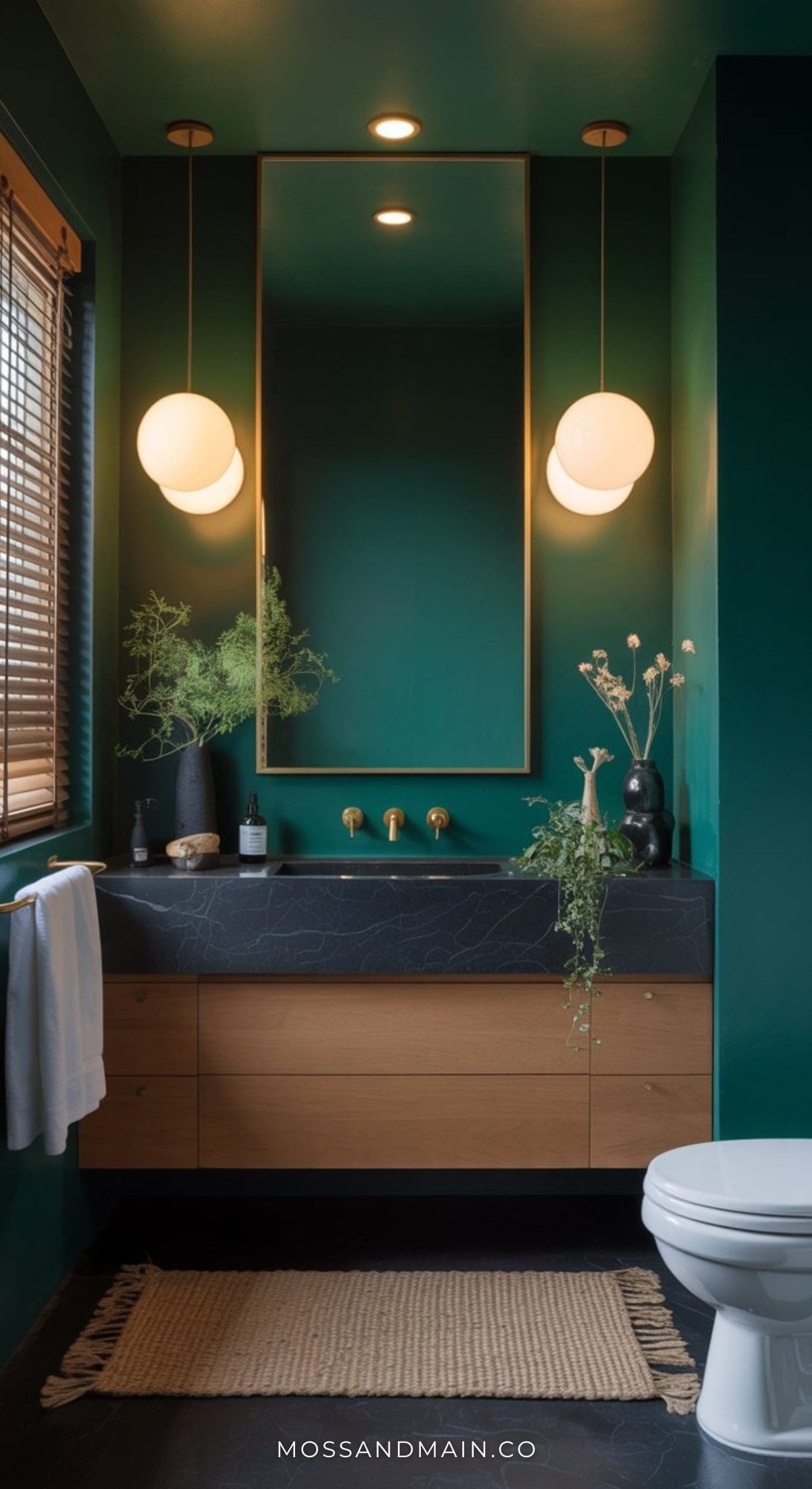 A moody bathroom with deep green walls, a large mirror, globe pendant lights, a wooden vanity with a black countertop, lush plants, and a beige woven rug. Sunlight filters through wooden blinds to complete this luxe bathroom retreat.