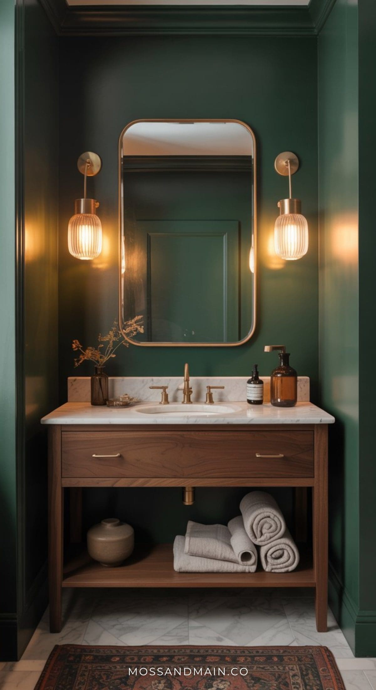 A moody bathroom with dark green walls, a wooden vanity topped with marble, a rectangular mirror, two brass wall sconces, and neatly folded towels. Dried flowers and luxe toiletries complete this sophisticated space.