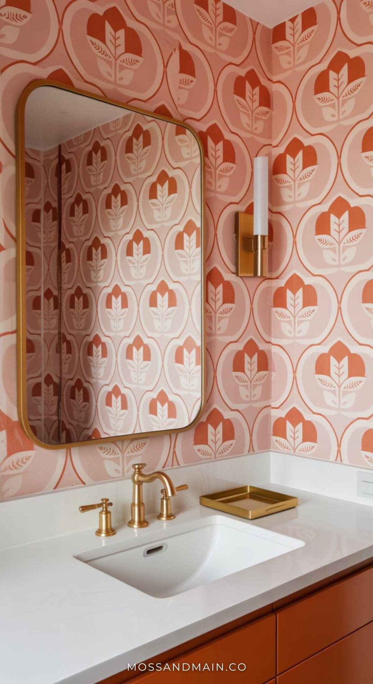 Modern and stylish pink bathroom with patterned pink and white floral wallpaper, a large rectangular mirror with rounded edges, brass faucet, matching light fixture, white countertop, and a decorative gold tray. Perfect for bold pink bathroom ideas.