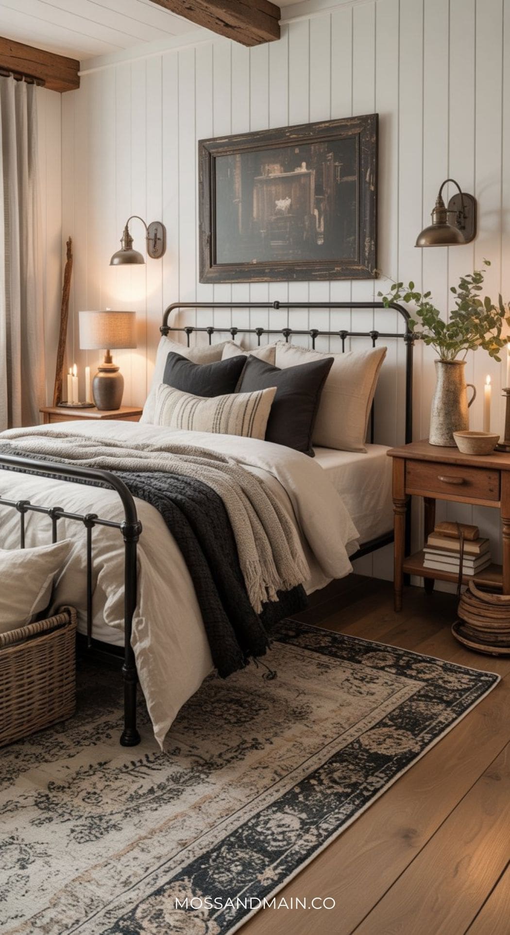 A cozy moody bedroom with modern farmhouse style features a black metal bed frame, layered neutral bedding, a vintage rug, wooden nightstands with decor, wall lamps, candles, and a framed art piece above the bed.