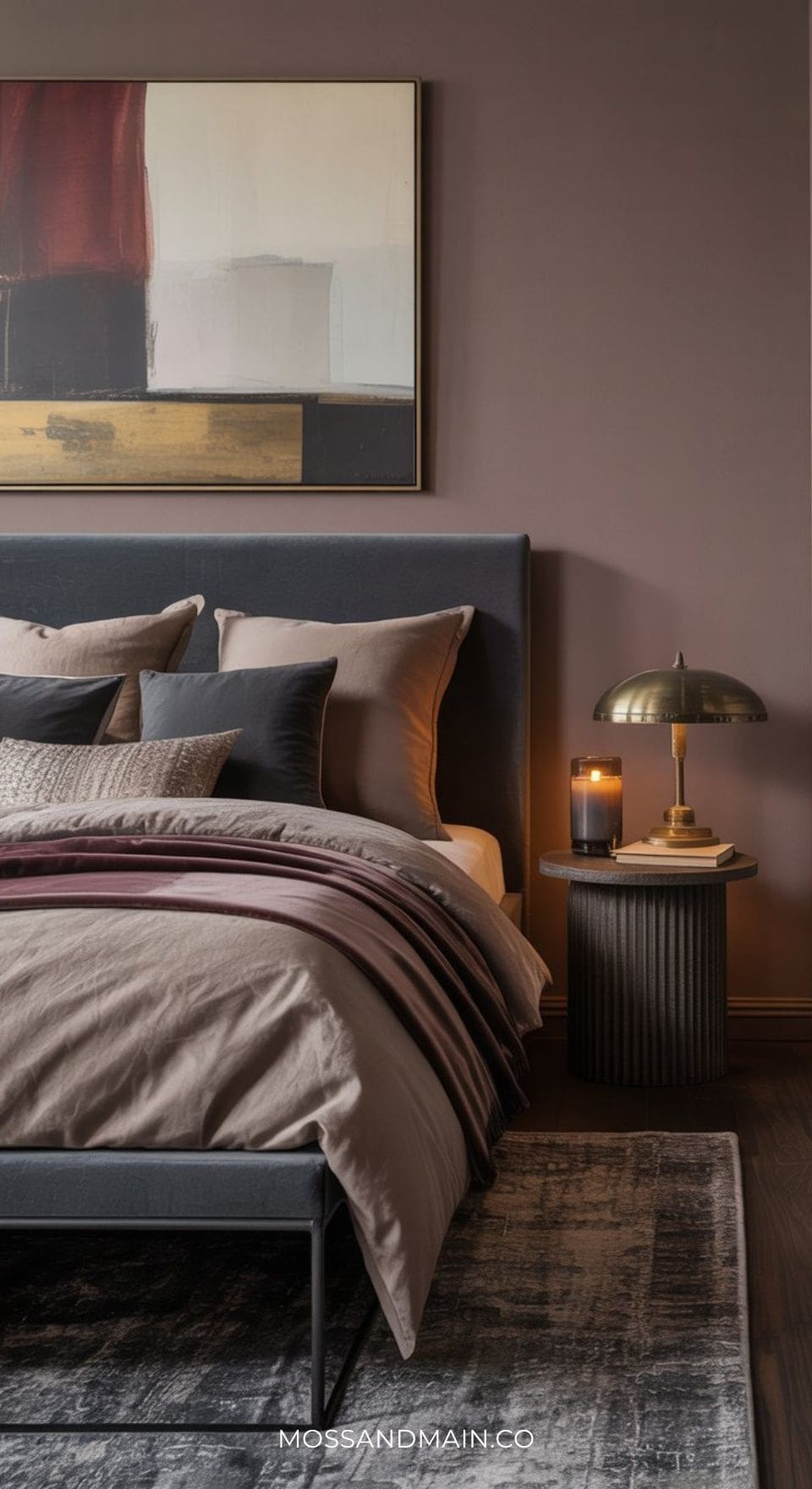 A modern grey bedroom featuring a grey headboard, taupe and burgundy bedding, an abstract painting above the bed, a gold lamp, candle, and book on a round nightstand, set against mauve walls and a patterned rug—perfect for stylish bedroom ideas.