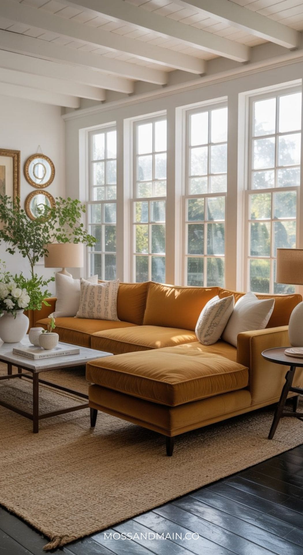 A bright living room with large windows, a mustard couch, white and patterned pillows, a coffee table with flowers, side tables with lamps, and a neutral rug on dark wood floors—perfect for those seeking mustard sofa living room ideas.