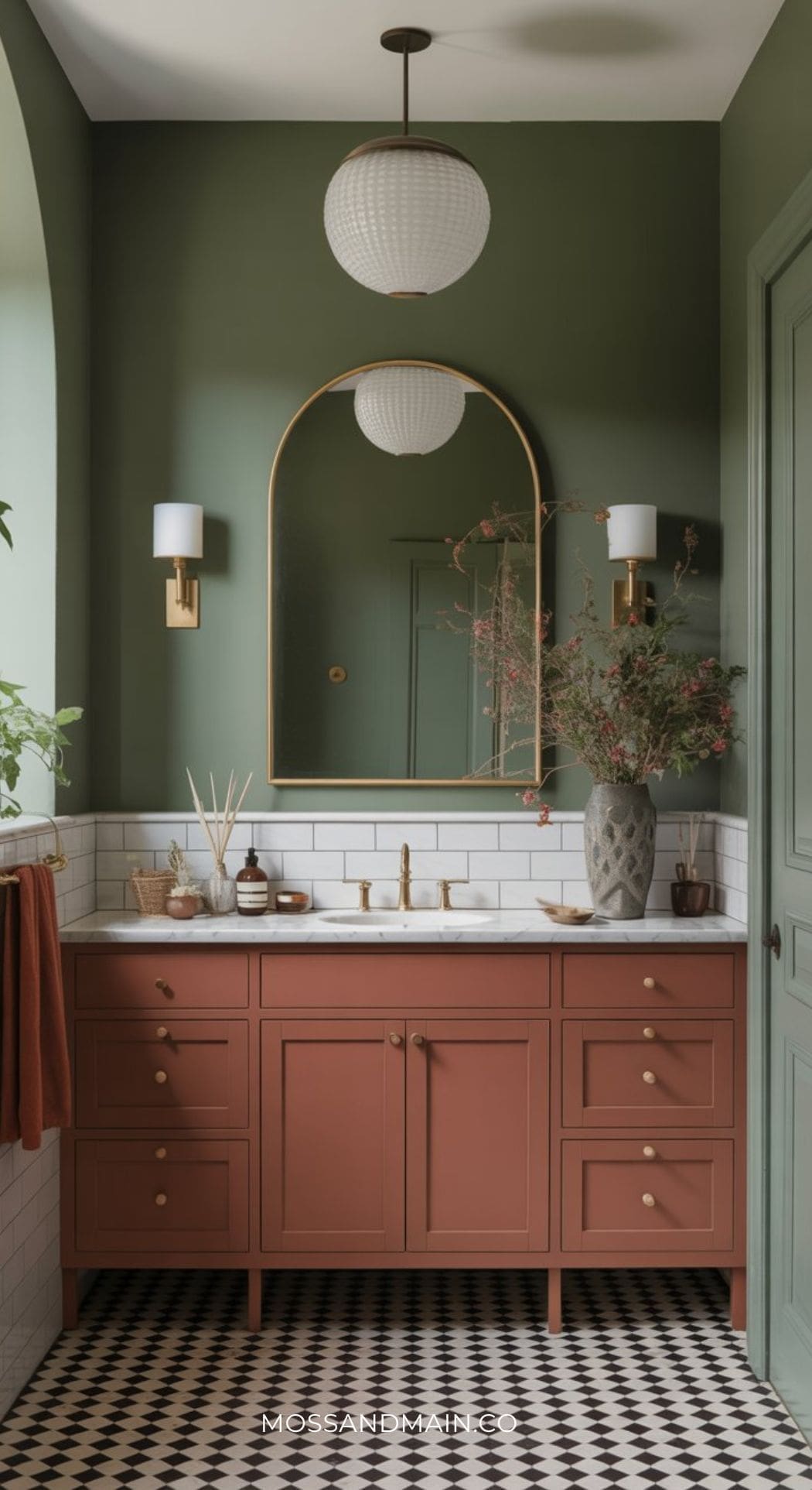 A stylish bathroom with sage green walls, a large arched mirror, globe pendant light, and a terracotta vanity with gold hardware. The white subway tile backsplash and decor complete the earthy color scheme.