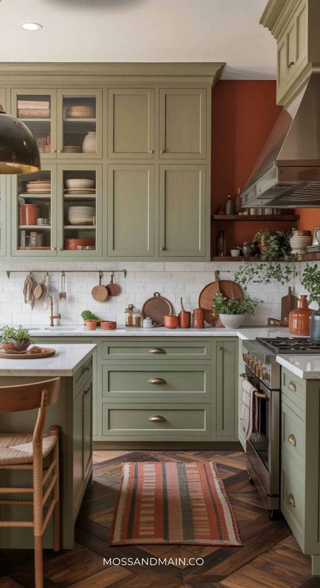 A cozy kitchen with a rust and sage color scheme features sage green cabinets, white countertops, a white tile backsplash, copper cookware, and terracotta accents. A striped rug and wooden chair add warmth, while open shelves display dishes and plants.