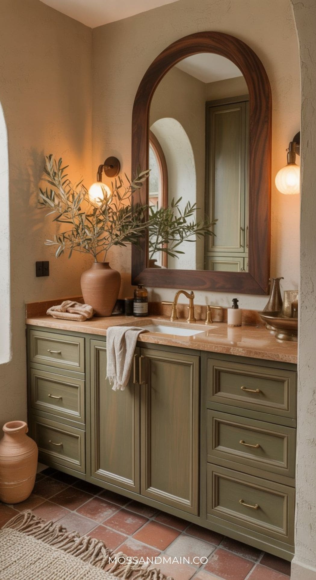A bathroom with sage green cabinets, a beige countertop, a large wooden-framed mirror, and brass fixtures. Warm lighting and earthy decor create a cozy, rustic atmosphere enhanced by a timeless color scheme.